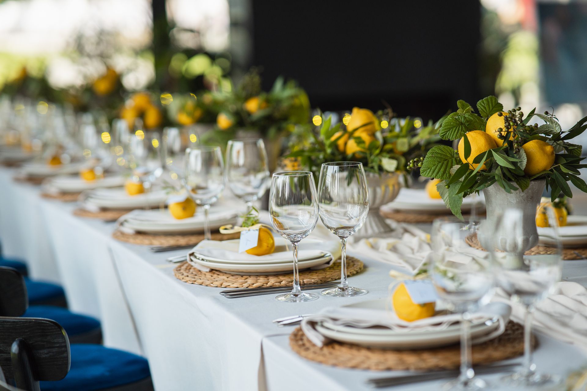 Long table set for a meal with yellow lemons as centerpieces and place settings; blue chairs.