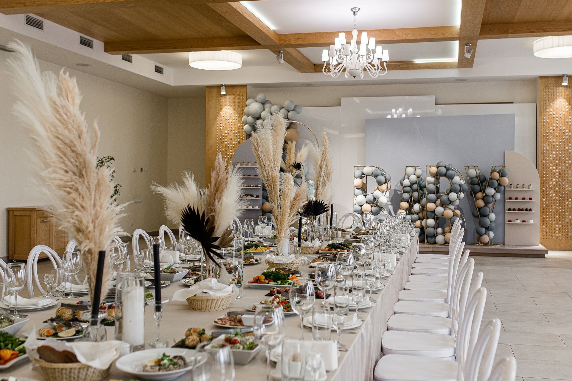 Long table set for a celebration, decorated with food, flowers, and balloons. White chairs and neutral decor.