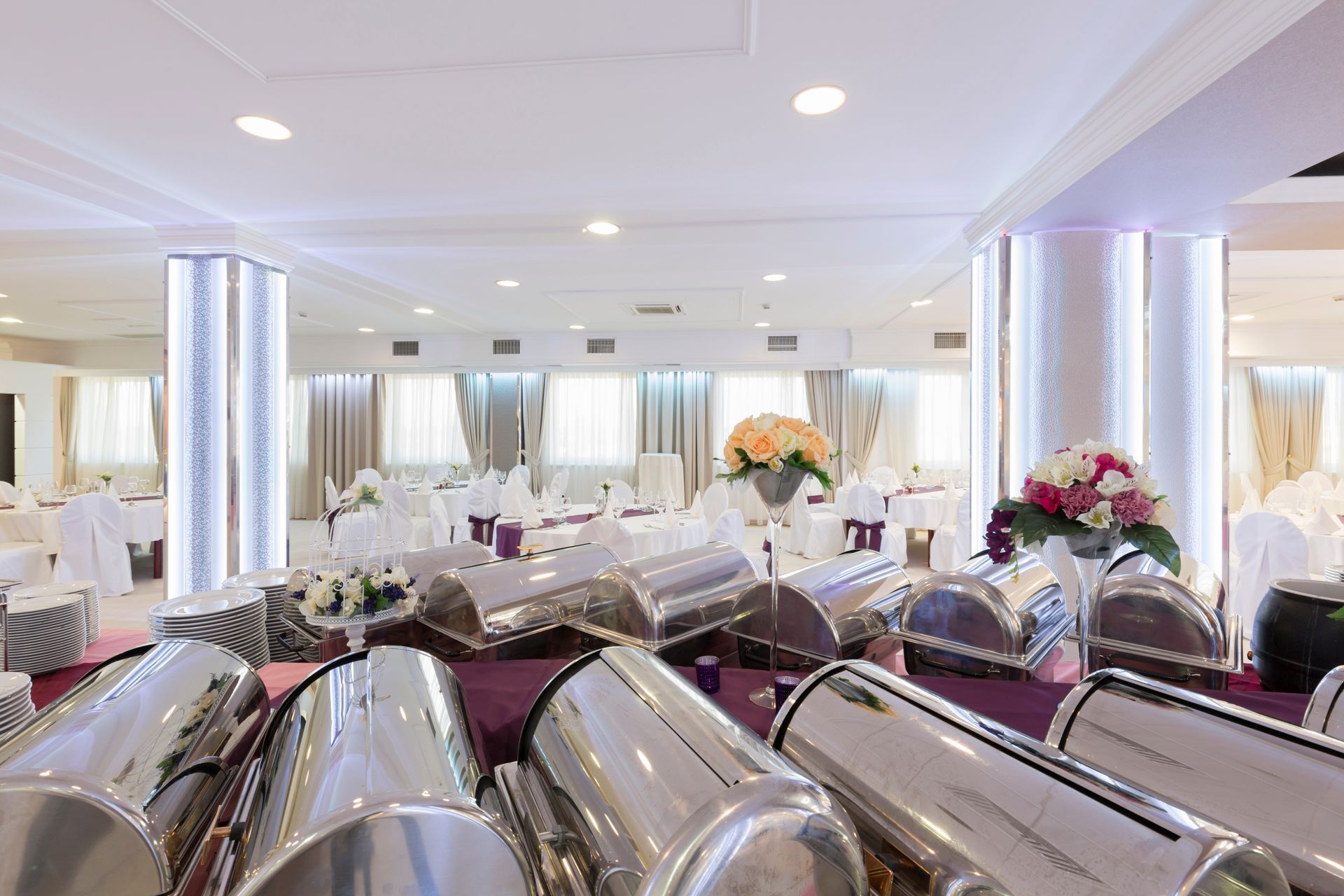 Catering buffet with silver chafing dishes in a formal dining hall with white tablecloths.
