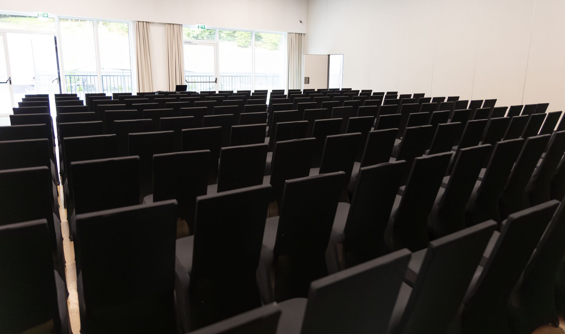 Rows of empty black chairs in a large, bright room with windows and a door.