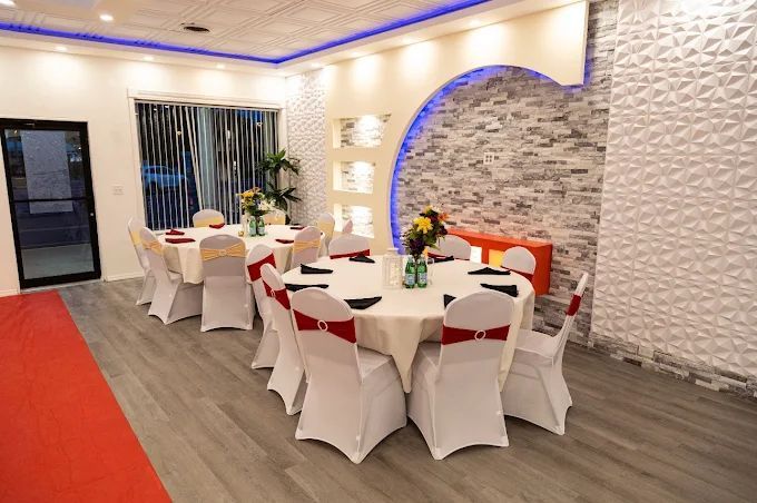 Interior of a banquet hall with round tables set for a celebration. Red carpet, white chairs, and decorative wall.
