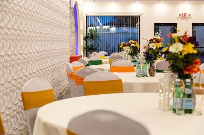Tables set for a celebration in a brightly lit room, featuring colorful flower arrangements and chair sashes.