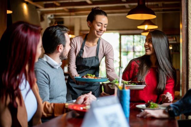 Waitress serving food to a group of people at a restaurant. Smiling, holding two plates; warm lighting.
