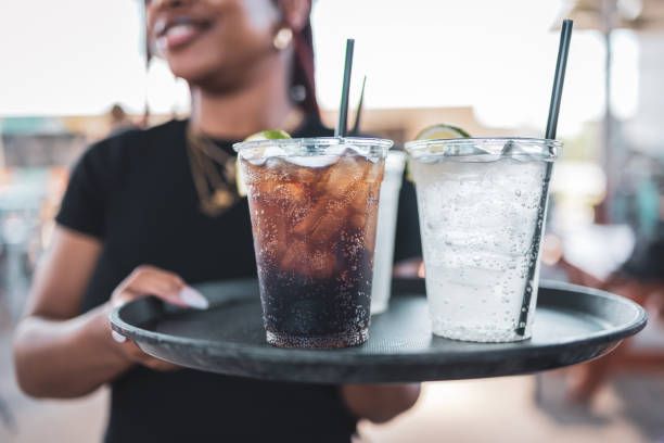Woman carrying a tray with three drinks: one cola, two clear, all with lime garnish and straws.