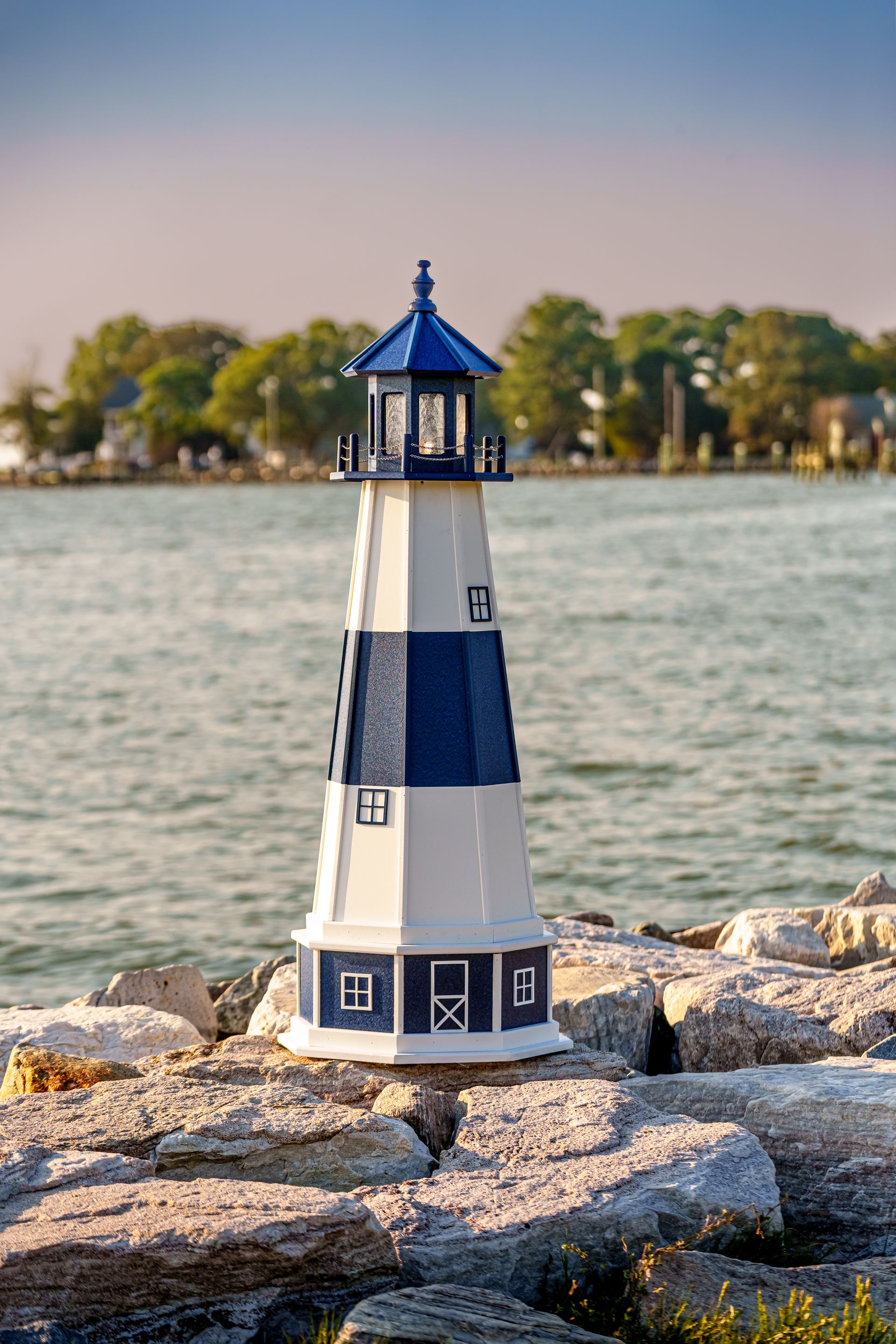 Brown and white lighthouse statue on a stone path, with a pond and trees in the background.