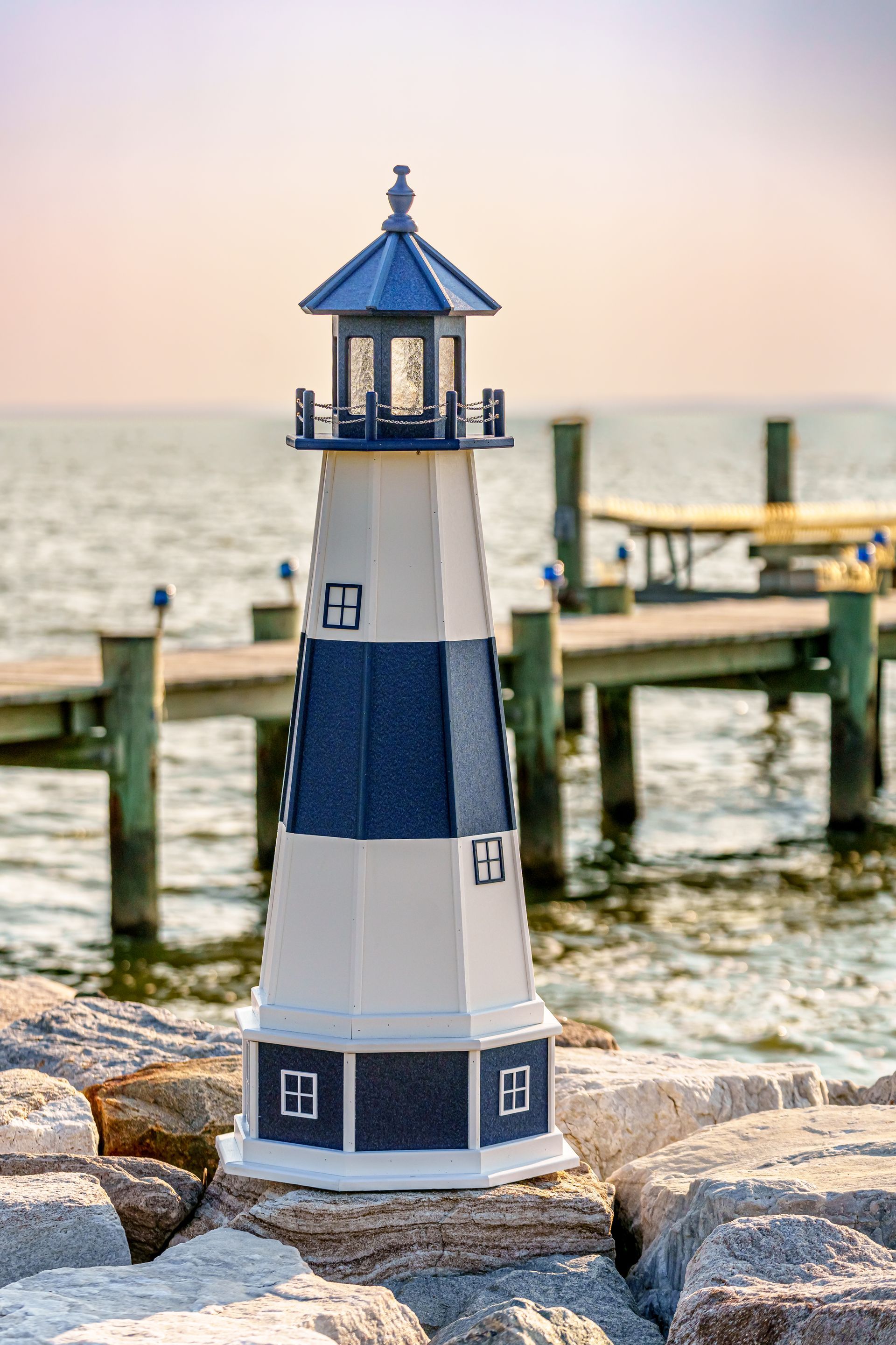 A decorative blue and white lighthouse model standing on rocky ground in front of a wooden dock and calm water.