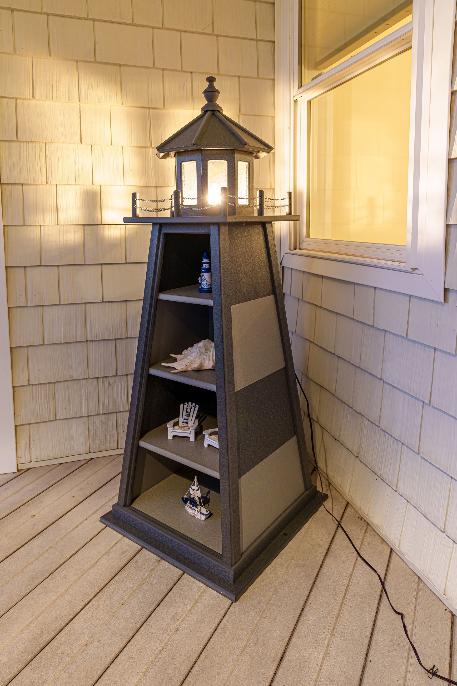 A gray and charcoal lighthouse-shaped display shelf stands on a porch, illuminated by a light at its top.