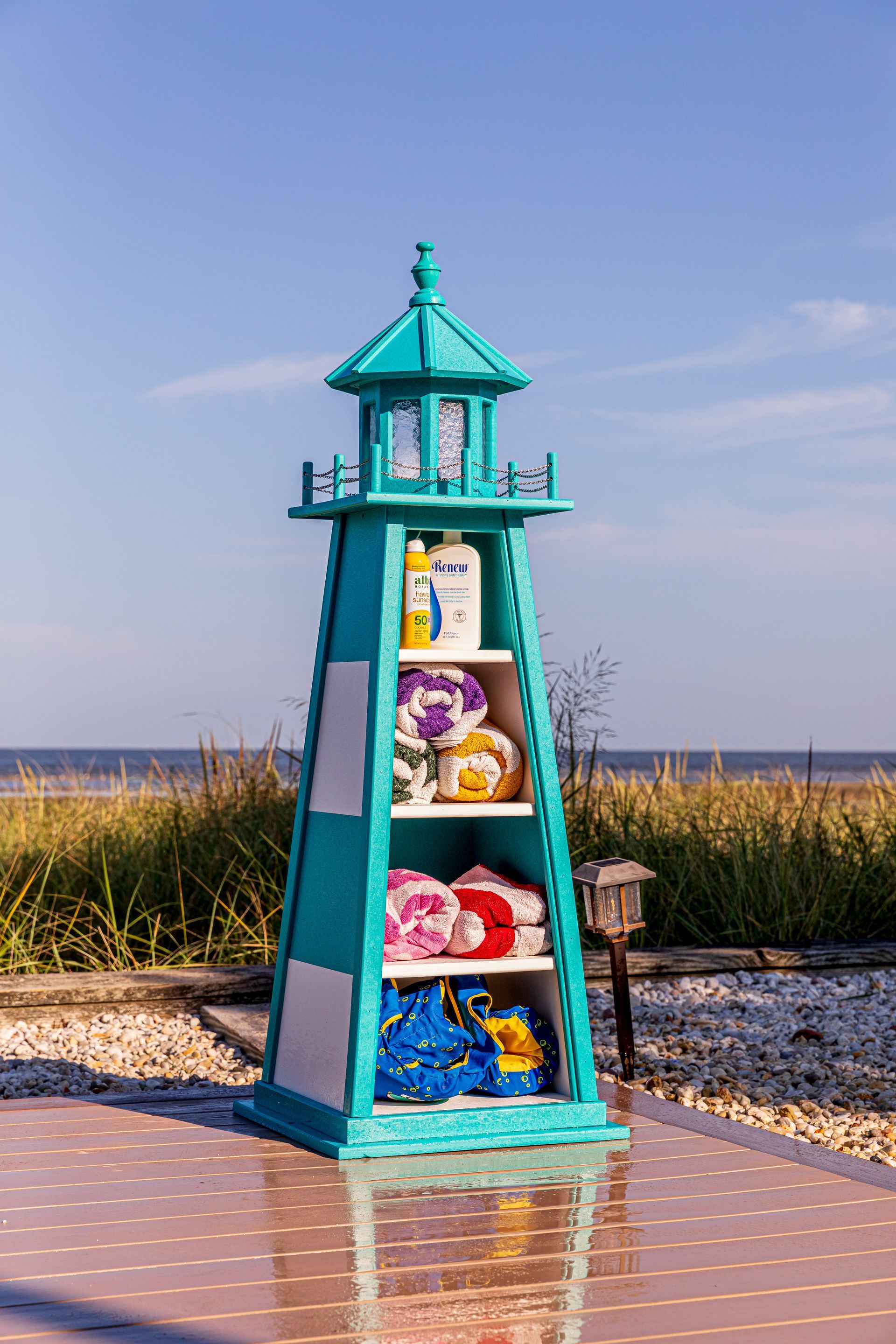 A teal lighthouse-shaped storage shelf holding bottles, rolled towels, and beach gear on a patio near a beach.