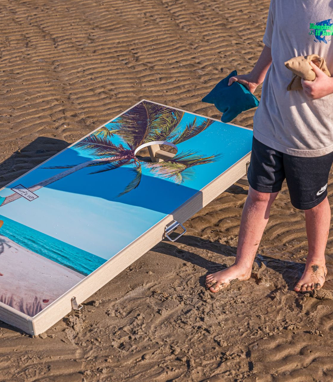 Person playing bean bag toss on beach. Board has palm tree scene. Blue bag in hand.