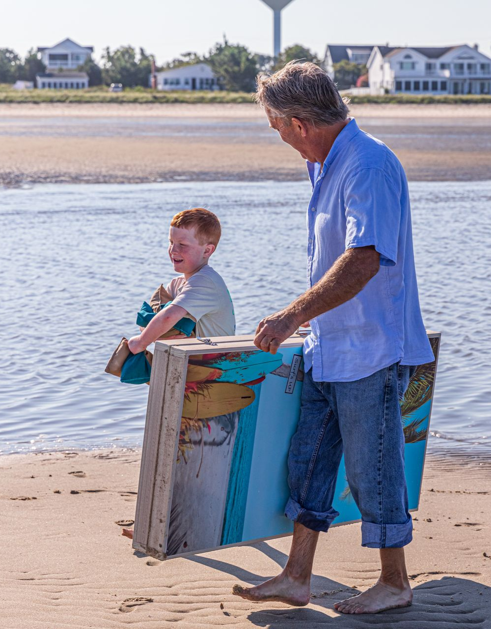 Man and child carry a colorful, weathered wooden structure on a beach near water.