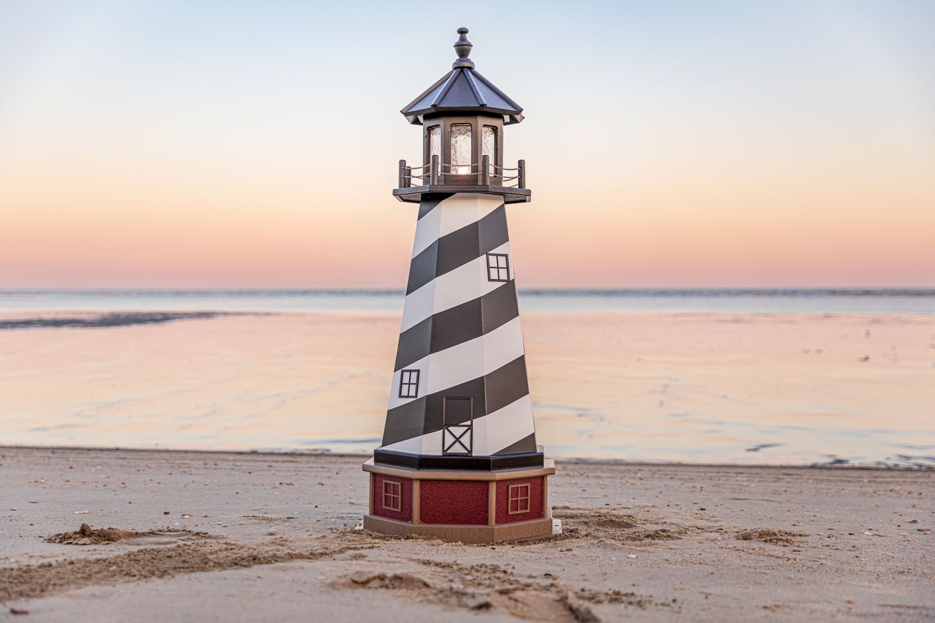 Model lighthouse with black and white stripes on a sandy beach at sunset.