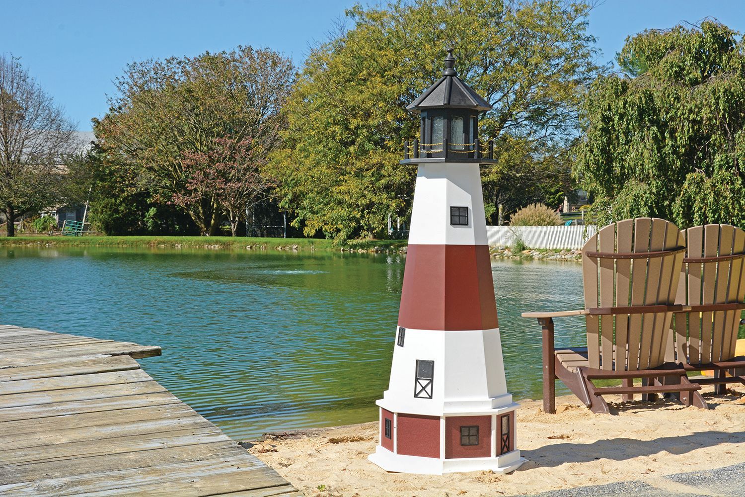 A red and white ornamental lighthouse stands on a sandy shore next to a wooden dock and two chairs by a calm pond.