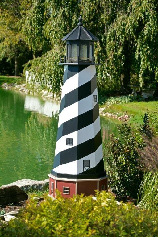 Decorative, black and white spiral-striped lighthouse model placed by a pond with trees in the background.