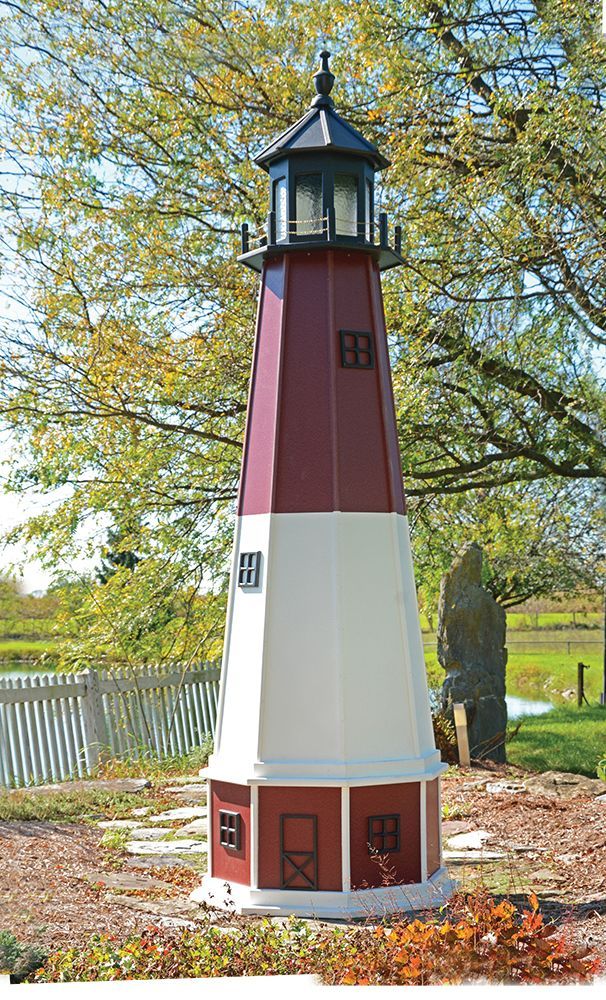 A red and white decorative lawn lighthouse in a garden setting with trees and a white picket fence in the background.