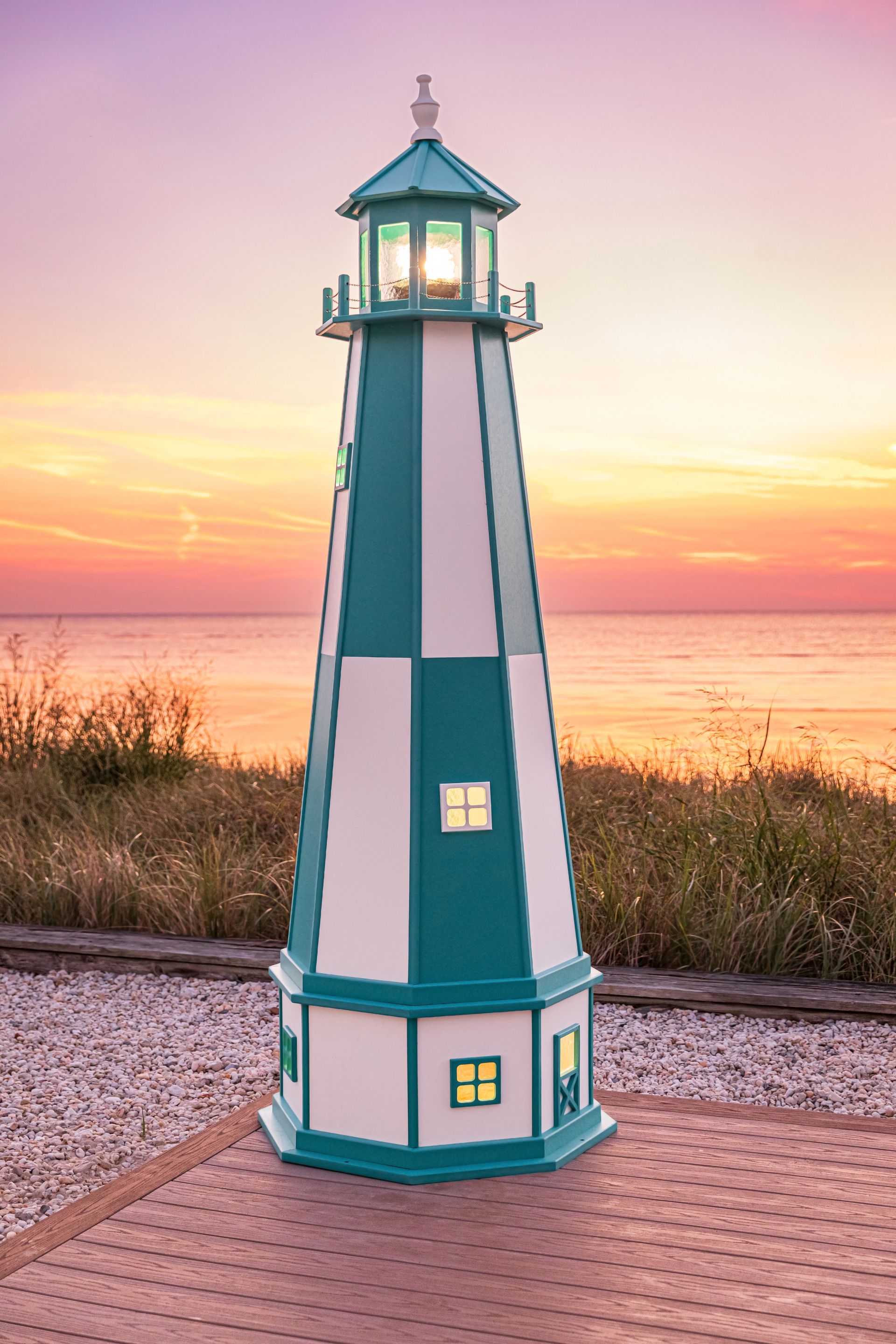 A teal and white decorative lighthouse standing on a wooden deck by the water at sunset.