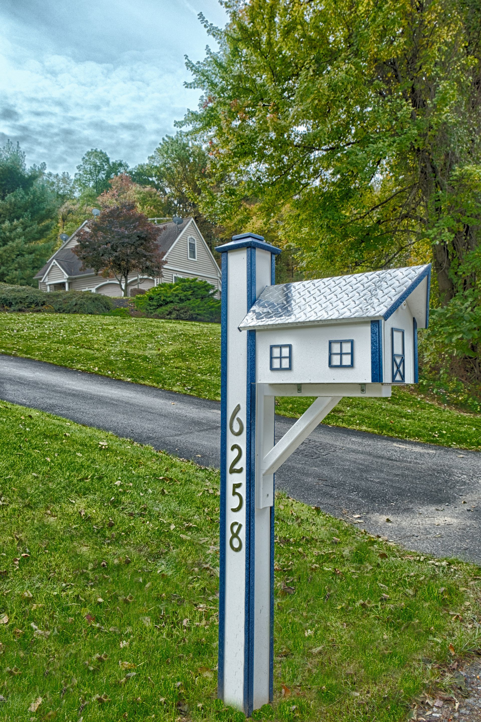 A white and blue mailbox shaped like a small house, displaying the address 6258, stands beside a driveway near a home.