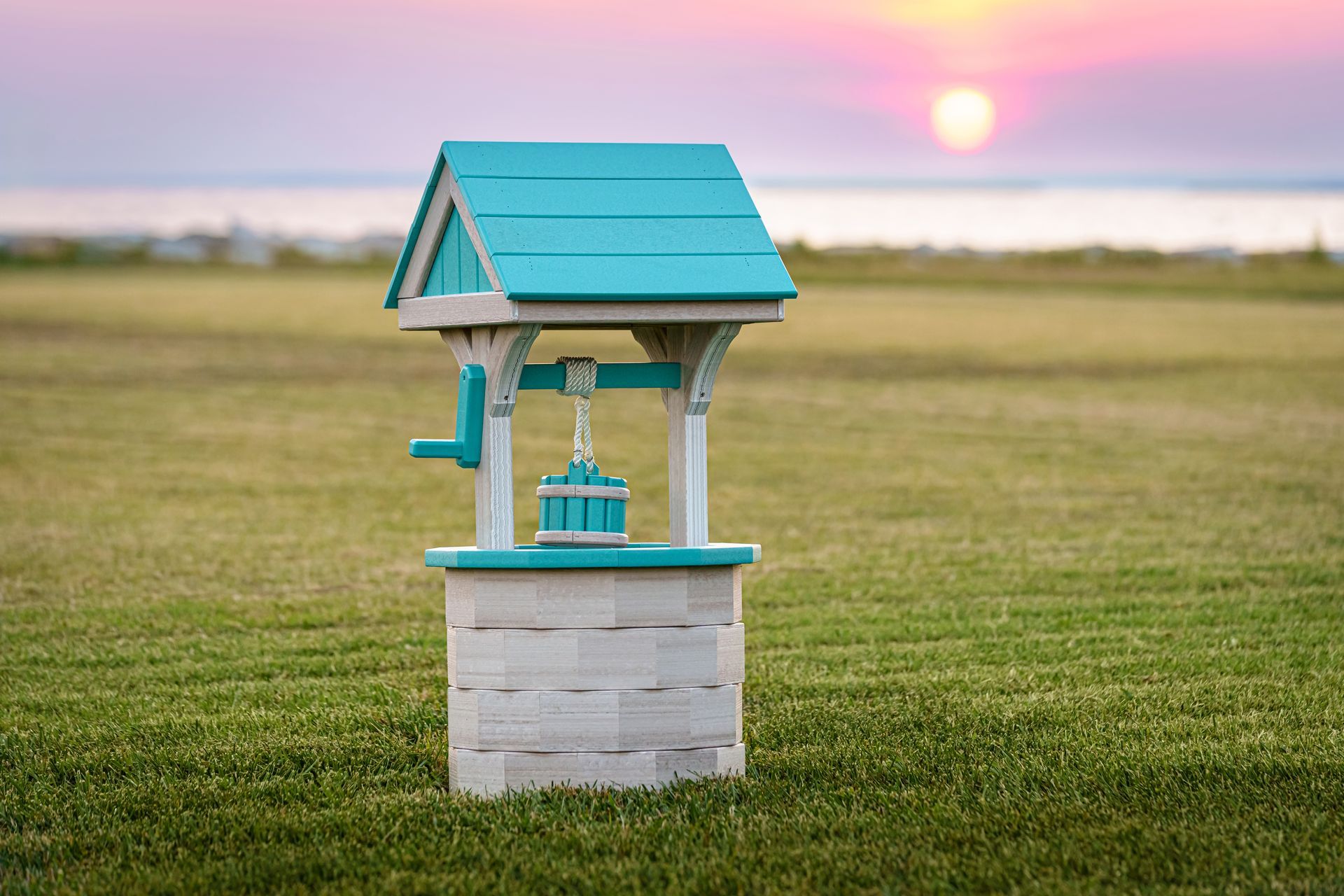 A decorative teal and white wishing well in a grassy field at sunset.