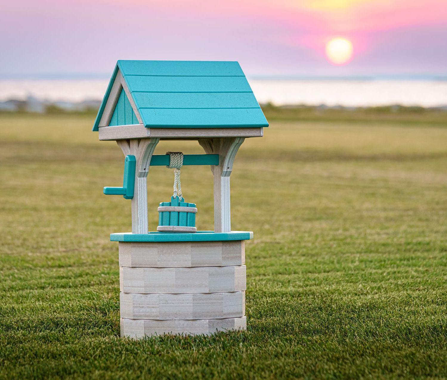 Decorative teal and white wishing well on a grassy field with a sunset in the background.