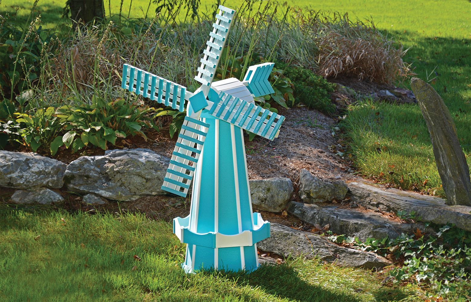 Blue and white decorative windmill in a grassy yard, with a stone wall and plants in the background.