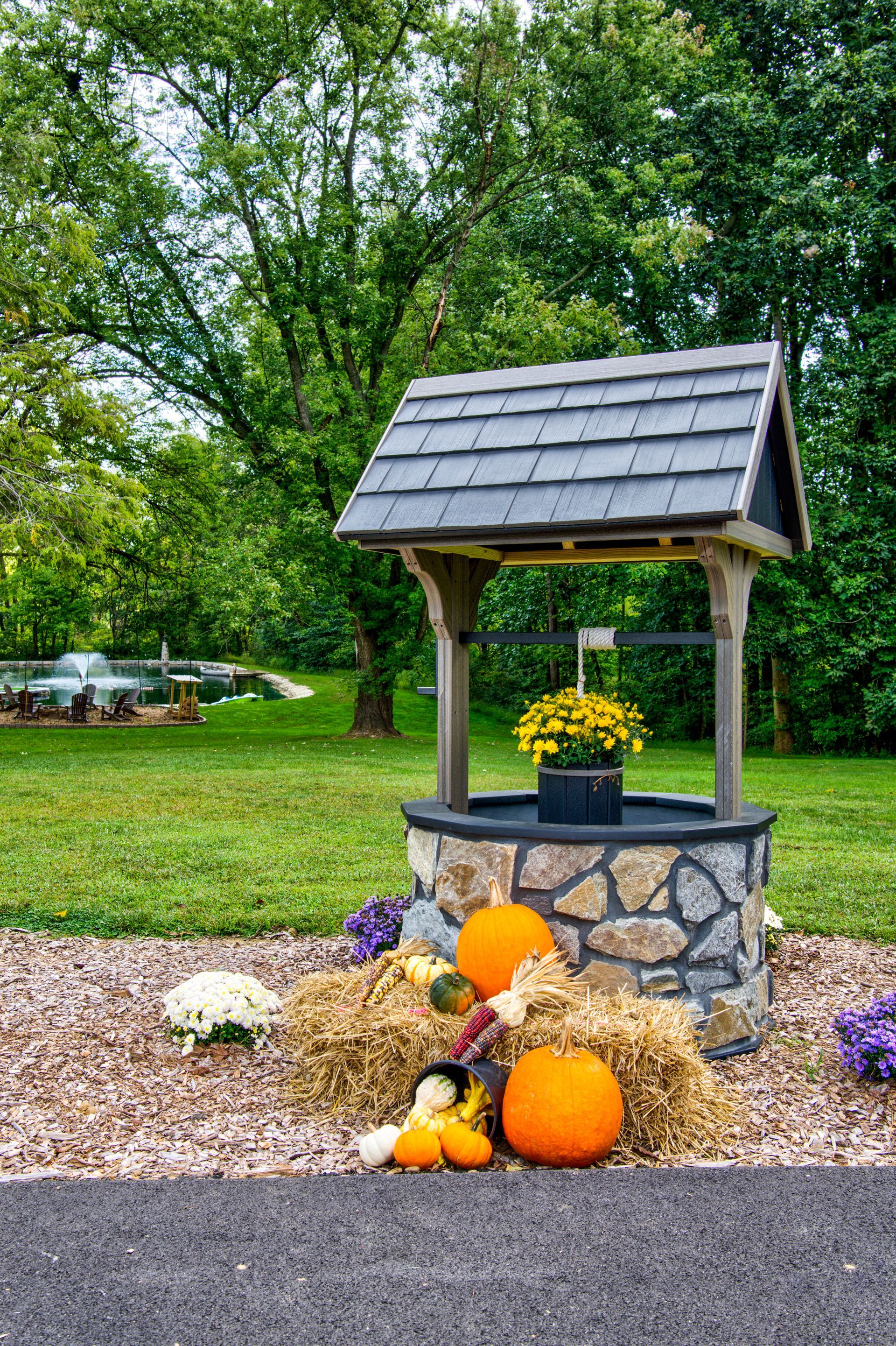A decorative stone wishing well decorated for autumn with pumpkins and mums on bales of hay in a grassy yard.