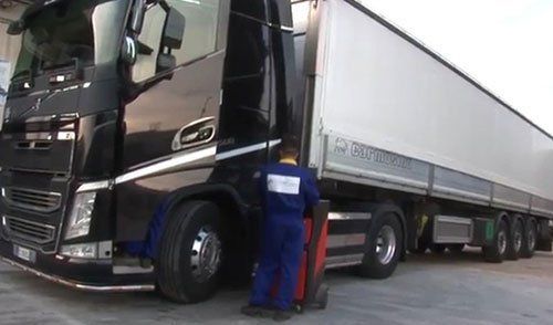 Un meccanico visto dal dietro con una tuta blu con un carrello con un macchinario rosso e accanto un camion nero con un rimorchio bianco