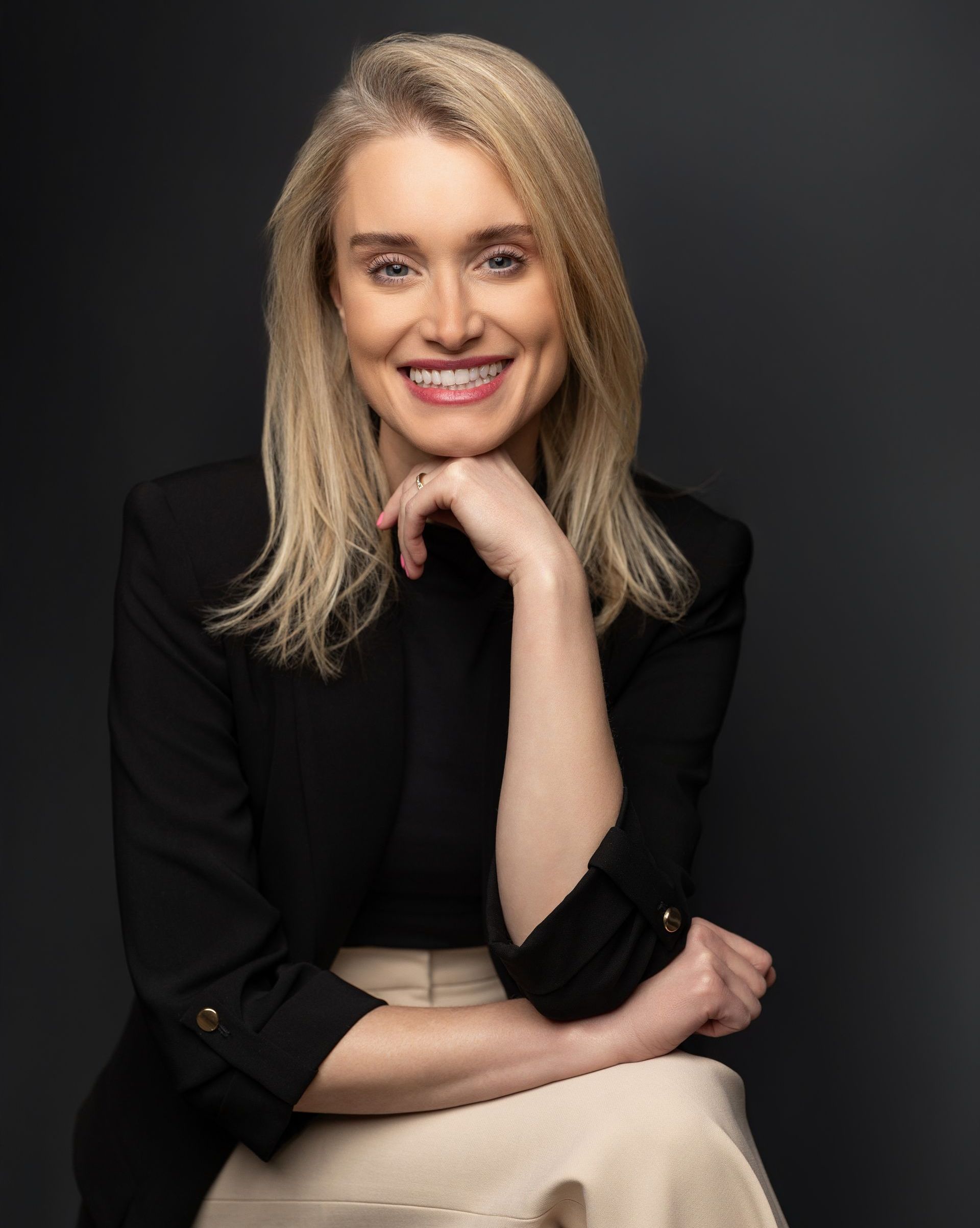 Blonde woman smiling, resting chin on hand, wearing black blazer and cream skirt, seated in front of a dark background.