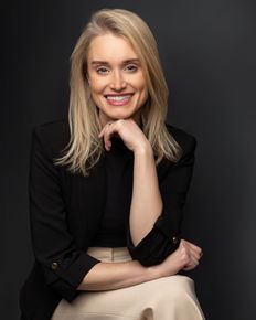 Blonde woman smiling, resting chin on hand, wearing black blazer and cream skirt, seated in front of a dark background.