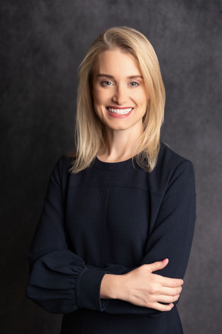 Blonde woman in navy dress smiles, arms crossed, dark gray background.