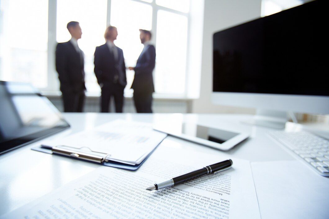 Office desk with pen, documents, computer, and blurred figures of three businesspeople.