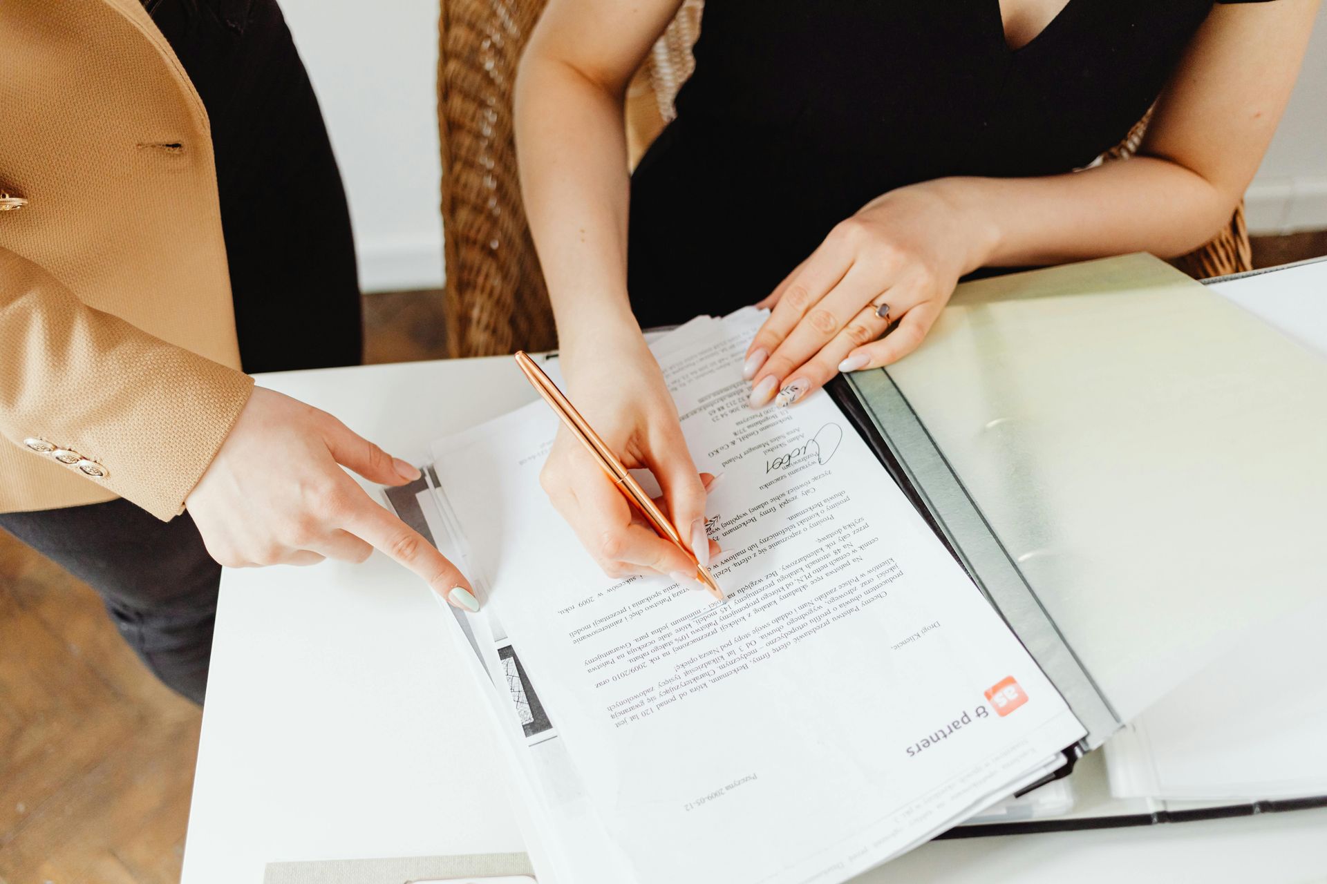 Woman signing a document, assisted by another person pointing. Table setting with neutral tones.