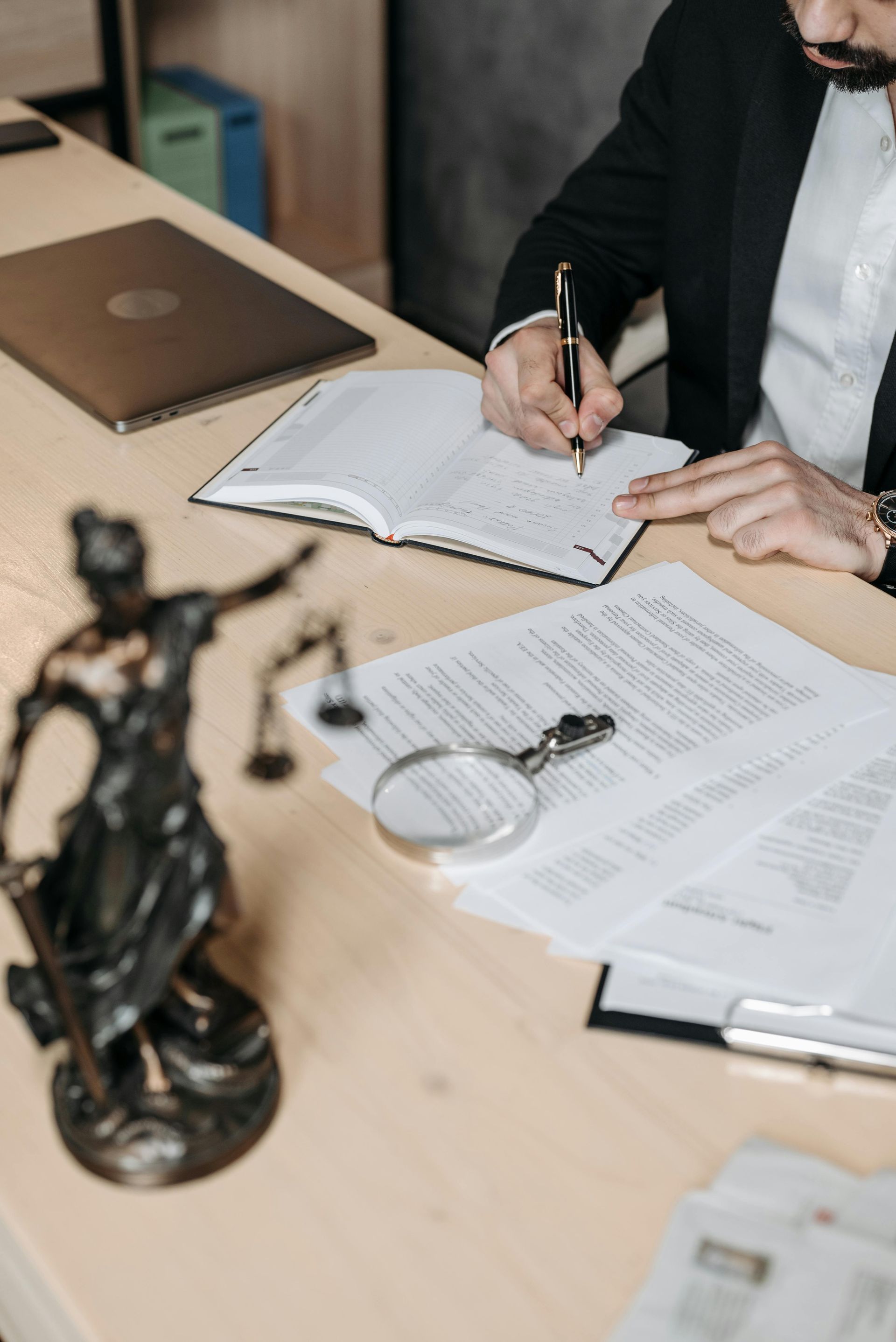 Person in suit writing on paper at desk with documents, statue of Lady Justice, and magnifying glass.