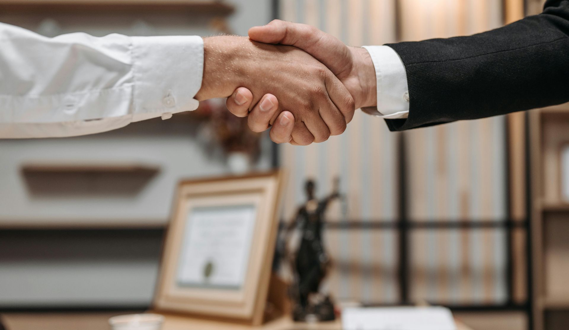Two people shaking hands, in an office. One in a white shirt, the other in a suit.