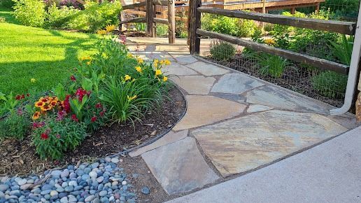 A stone walkway in a garden with flowers and a wooden fence.