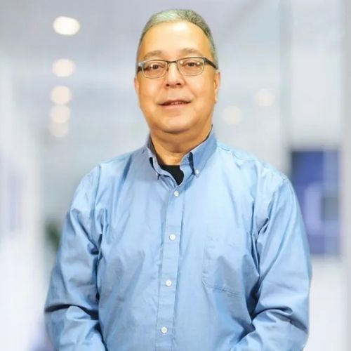 A man wearing glasses and a blue shirt is standing in a hallway.