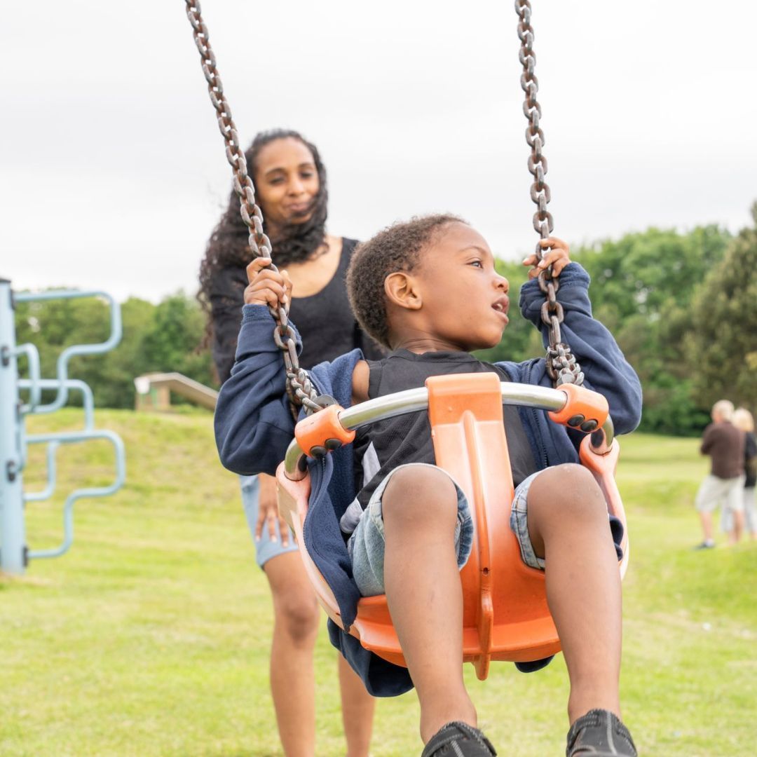 A boy is sitting on a swing with a woman standing behind him.