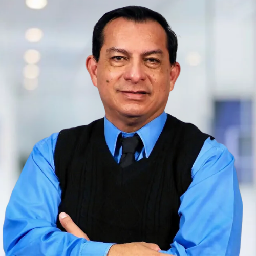 A man in a blue shirt and black vest stands with his arms crossed