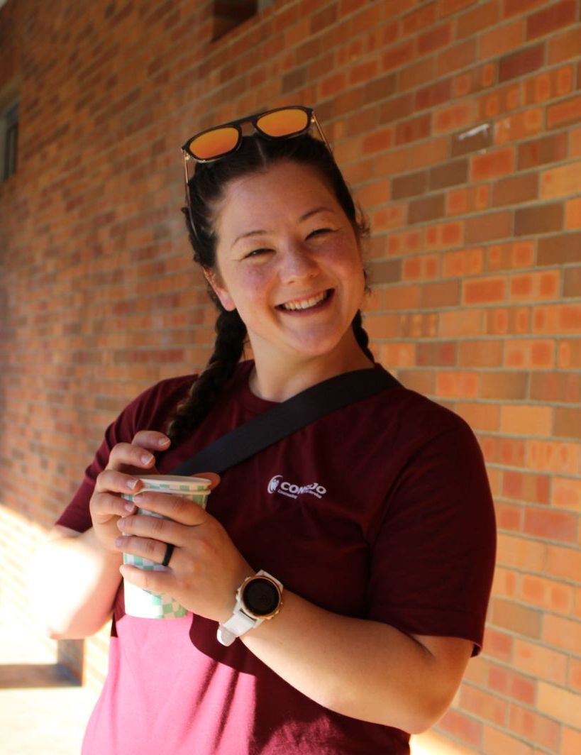 A woman wearing sunglasses and a maroon shirt is smiling and holding a cup