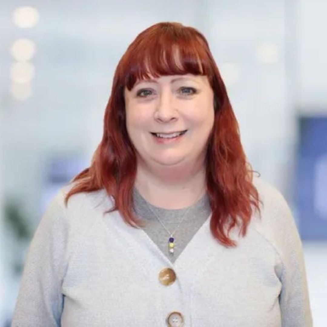 A woman with red hair is wearing a grey sweater and smiling for the camera.