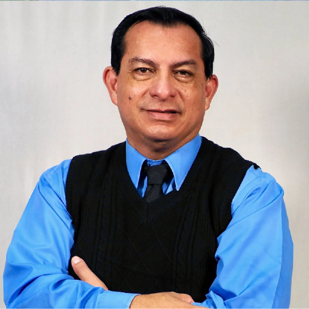 A man in a blue shirt and black vest stands with his arms crossed