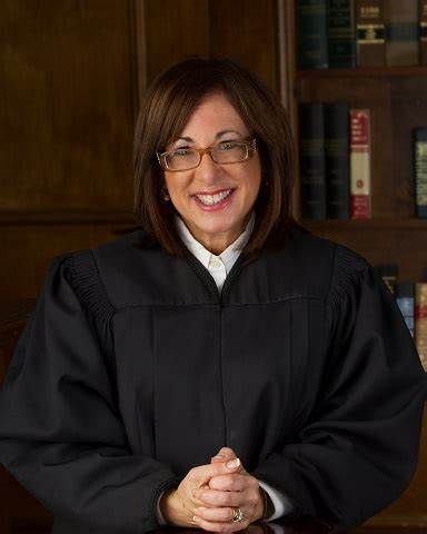 A female judge is smiling while sitting at a desk in front of a bookshelf.