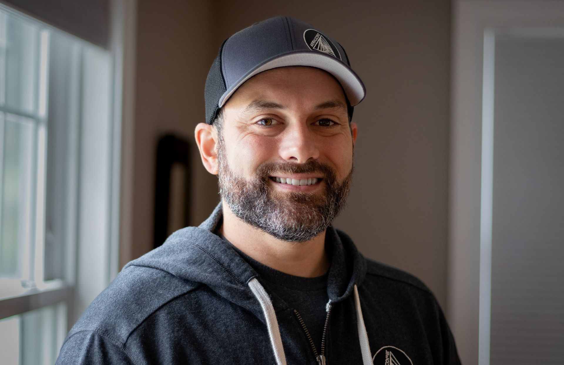 A man with a beard wearing a hat and a hoodie is smiling for the camera.