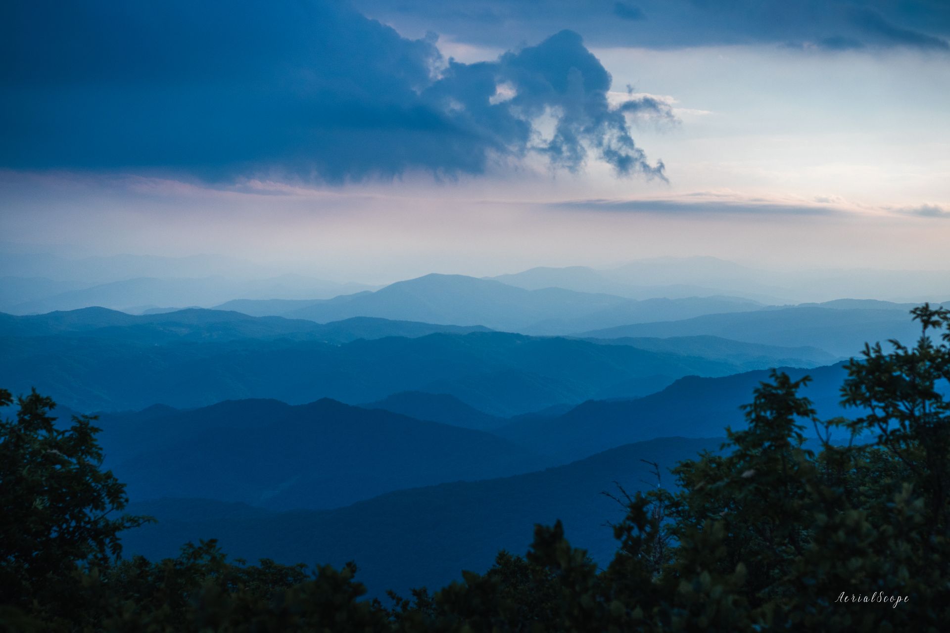 A view of a mountain range at sunset with a cloudy sky and trees in the foreground.