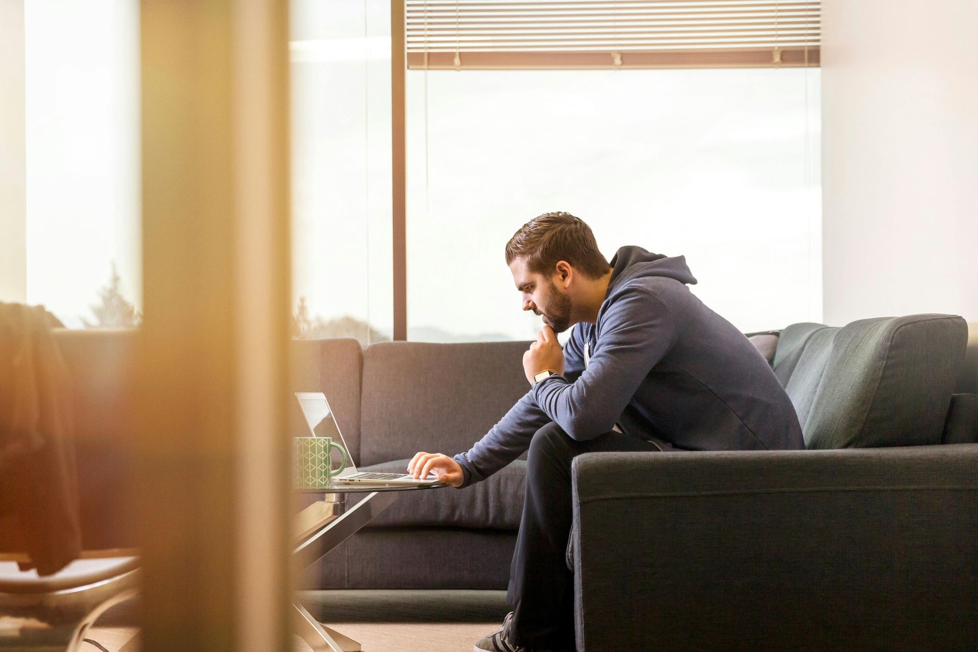 Man sitting on a couch looking at laptop, hand on chin. Window in background.