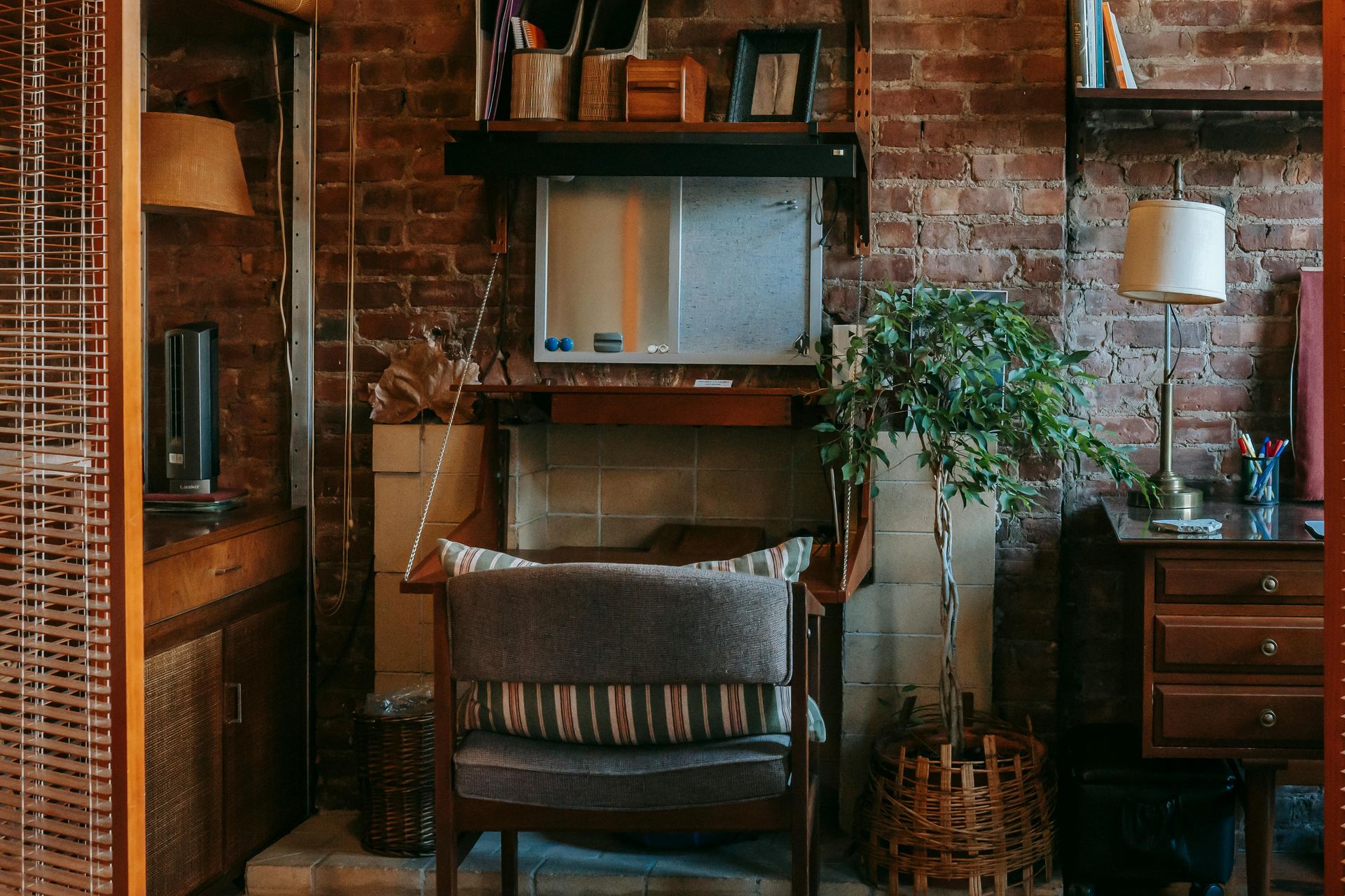 Cozy living area with brick wall. An armchair sits before a fireplace, flanked by a desk and cabinet, and a potted plant.