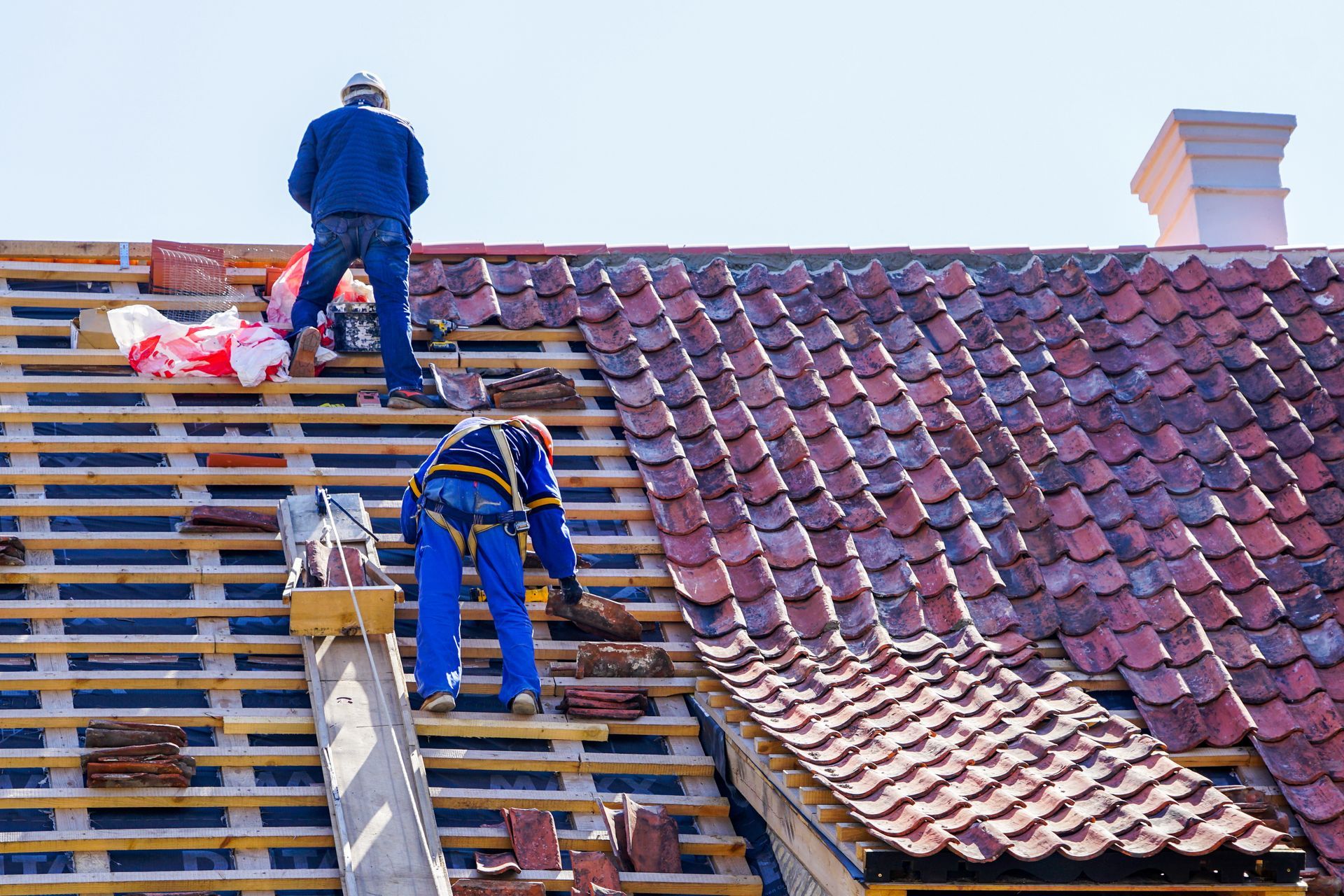 Two workers installing clay roof tiles on a sloped roof with exposed wooden battens.