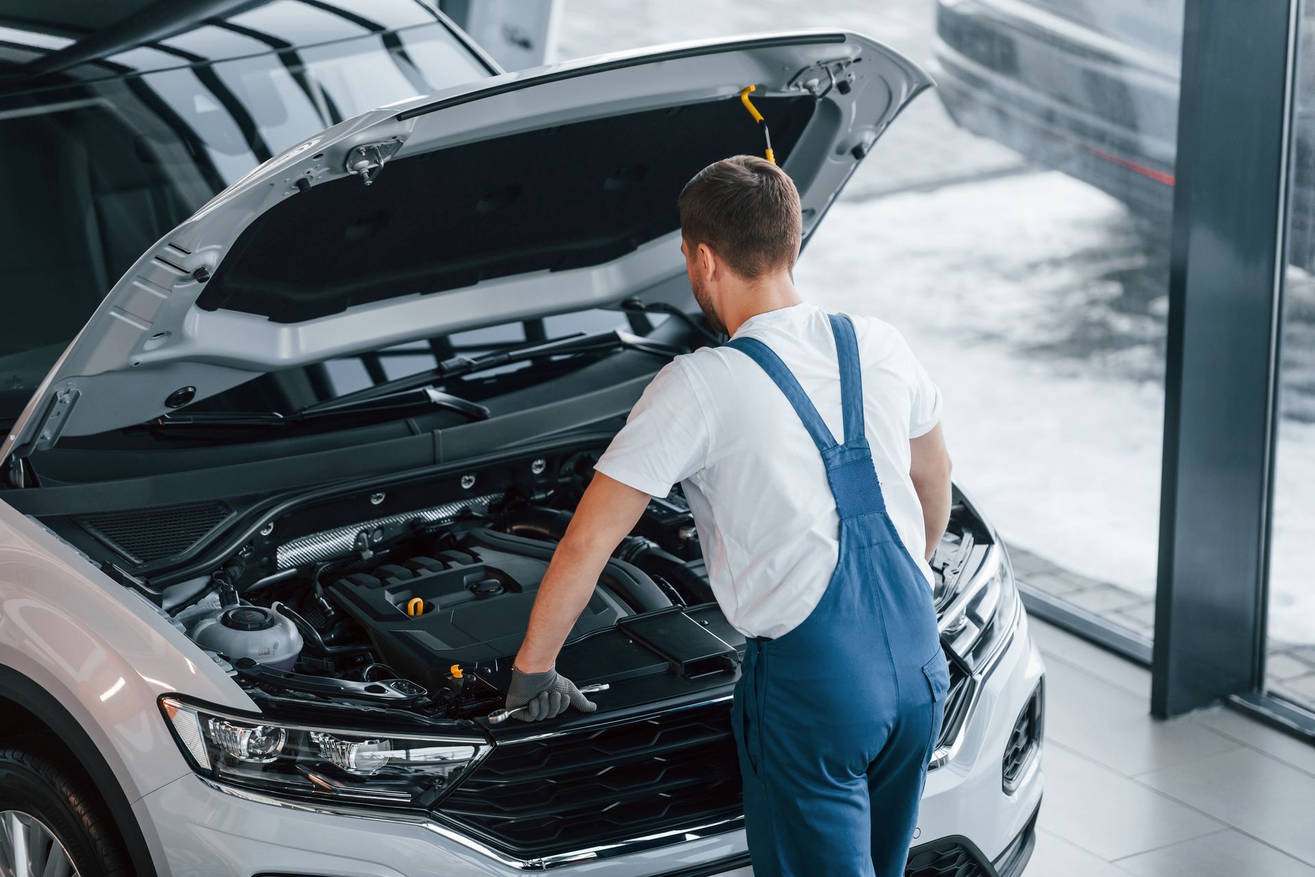 Hood is opened. Young man in white shirt and blue uniform repairs automobile.