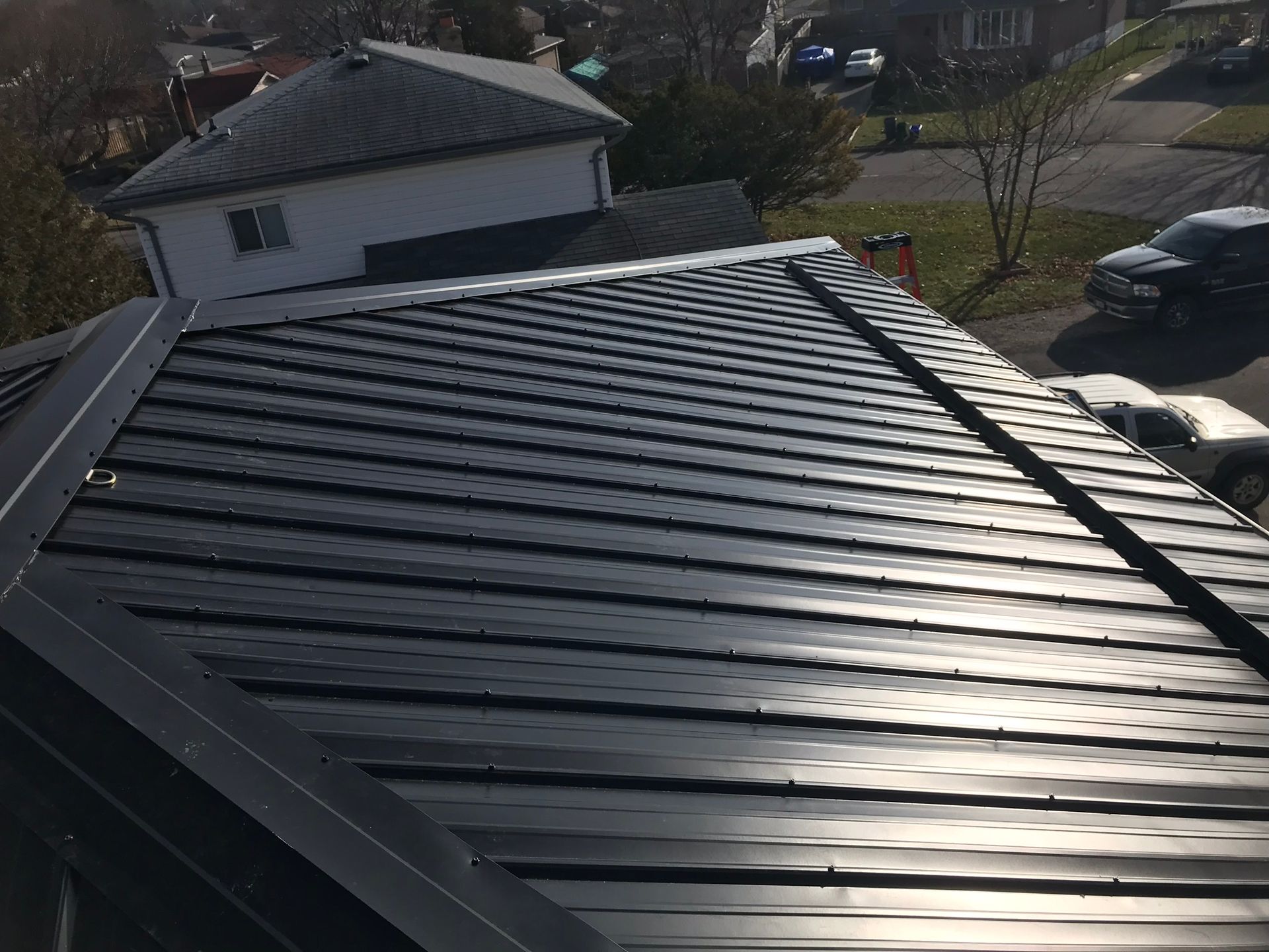 Black metal roof on a house in sunlight, viewed from above with a yard and parked car in the background