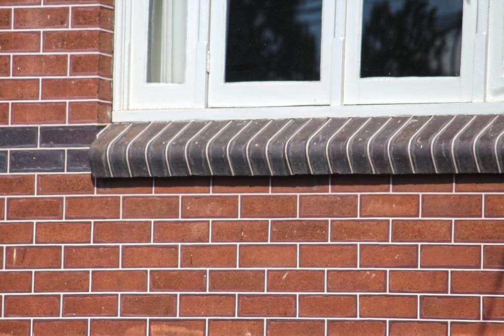Red brick wall with a white-framed window. A row of curved, dark bricks forms a window sill.