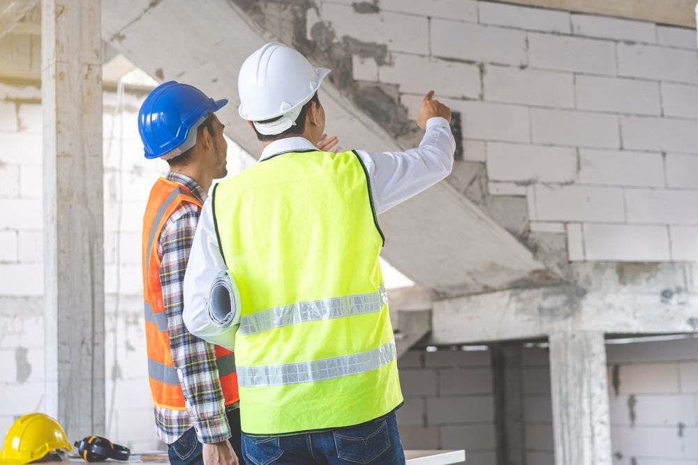 Two construction workers in safety vests and hard hats inspecting a building's interior. One points upward.