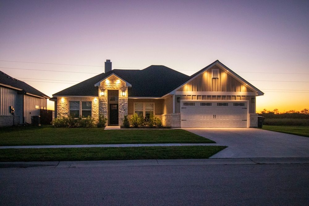 Single-story house with stone facade, lit up at dusk. Green lawn, driveway, and sunset in the background.